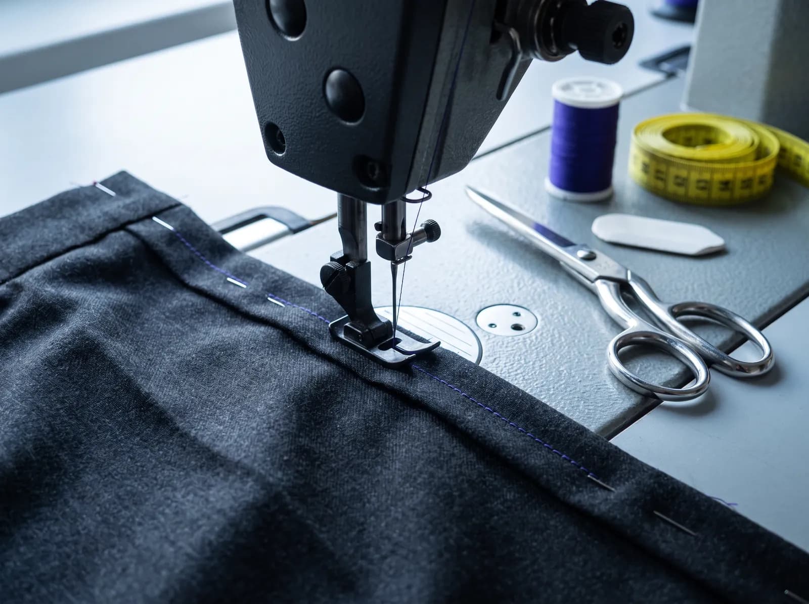 Symbolbild: Ein Hosensaum wird an einer Industrienähmaschine genäht, daneben Schere, Maßband und Garn.